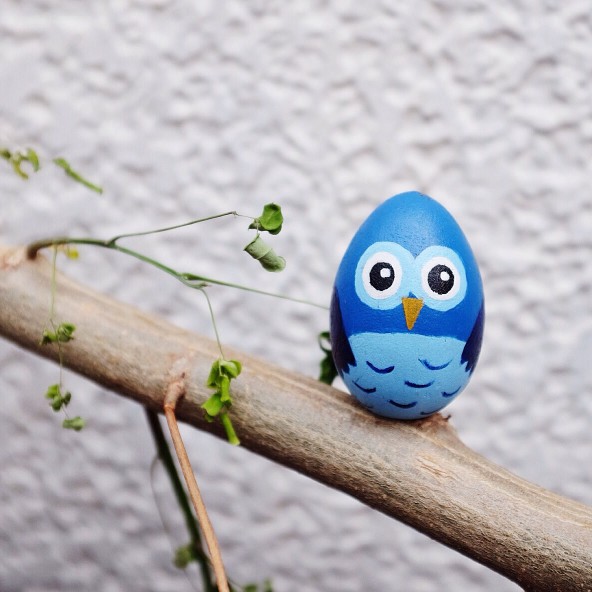 Blue egg decorated as an owl perched on a branch with small green leaves.