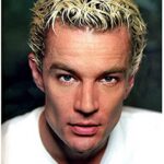 Man with spiky blond hair in a white shirt stares seriously at the camera.