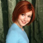 Smiling woman with short red hair in a blue top glances over her shoulder, set against a green background.