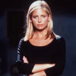 Blonde woman in a black top stands with arms crossed, gazing seriously at the camera against a dark backdrop.