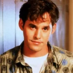 Young man with dark hair in a patterned shirt stands before lockers, looking ahead with a neutral expression.