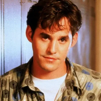 Young man with dark hair in a patterned shirt stands before lockers, looking ahead with a neutral expression.