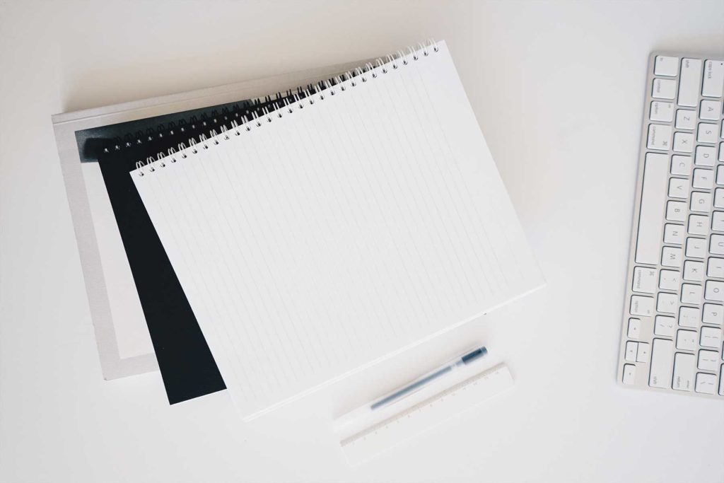 A lined notebook, pen, ruler, and keyboard neatly arranged on a white desk.