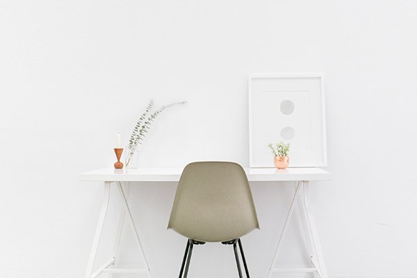 White desk with a plant, candle, framed artwork, and a simple gray chair placed in front.